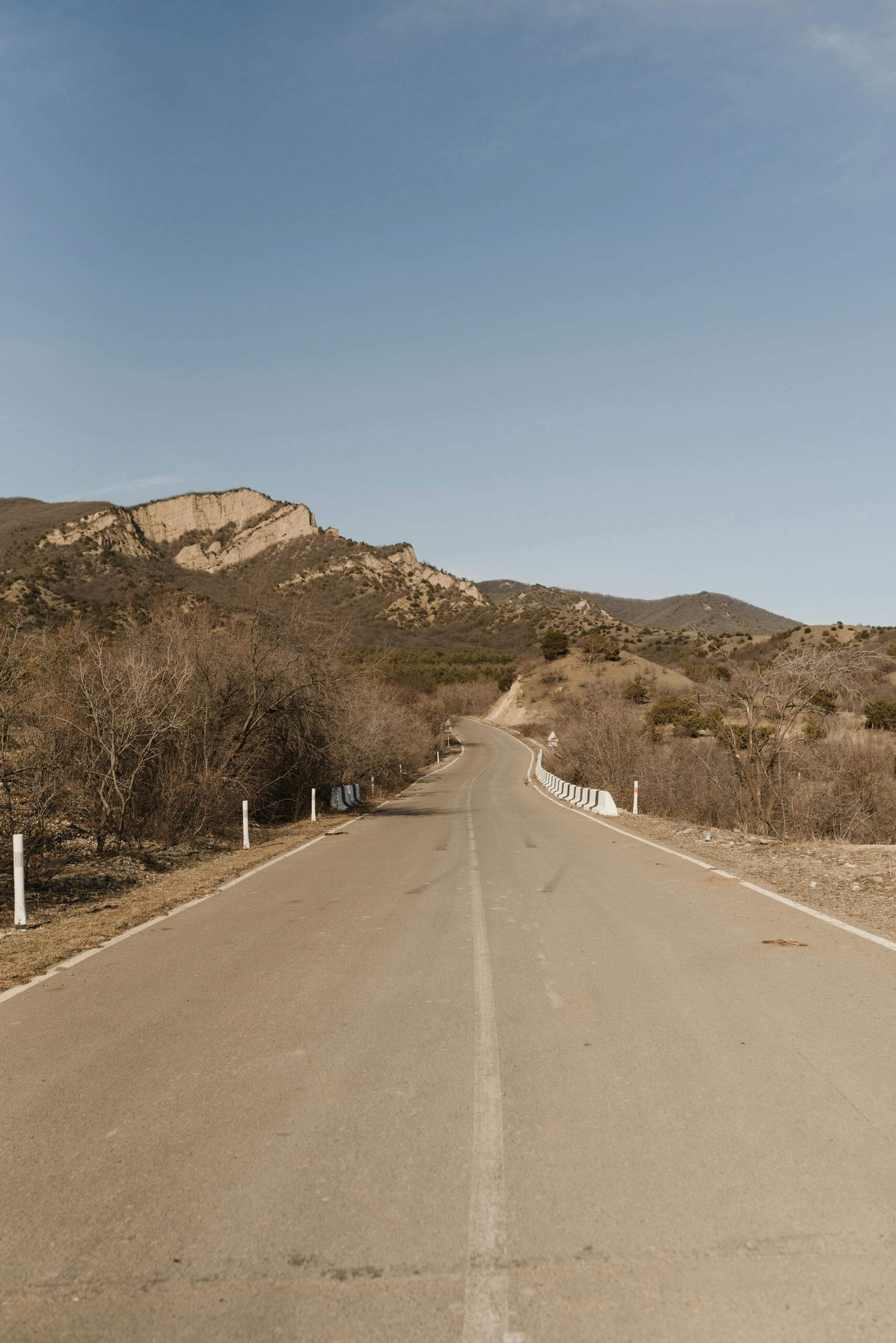 Empty rural road leading towards majestic mountains under a clear blue sky.