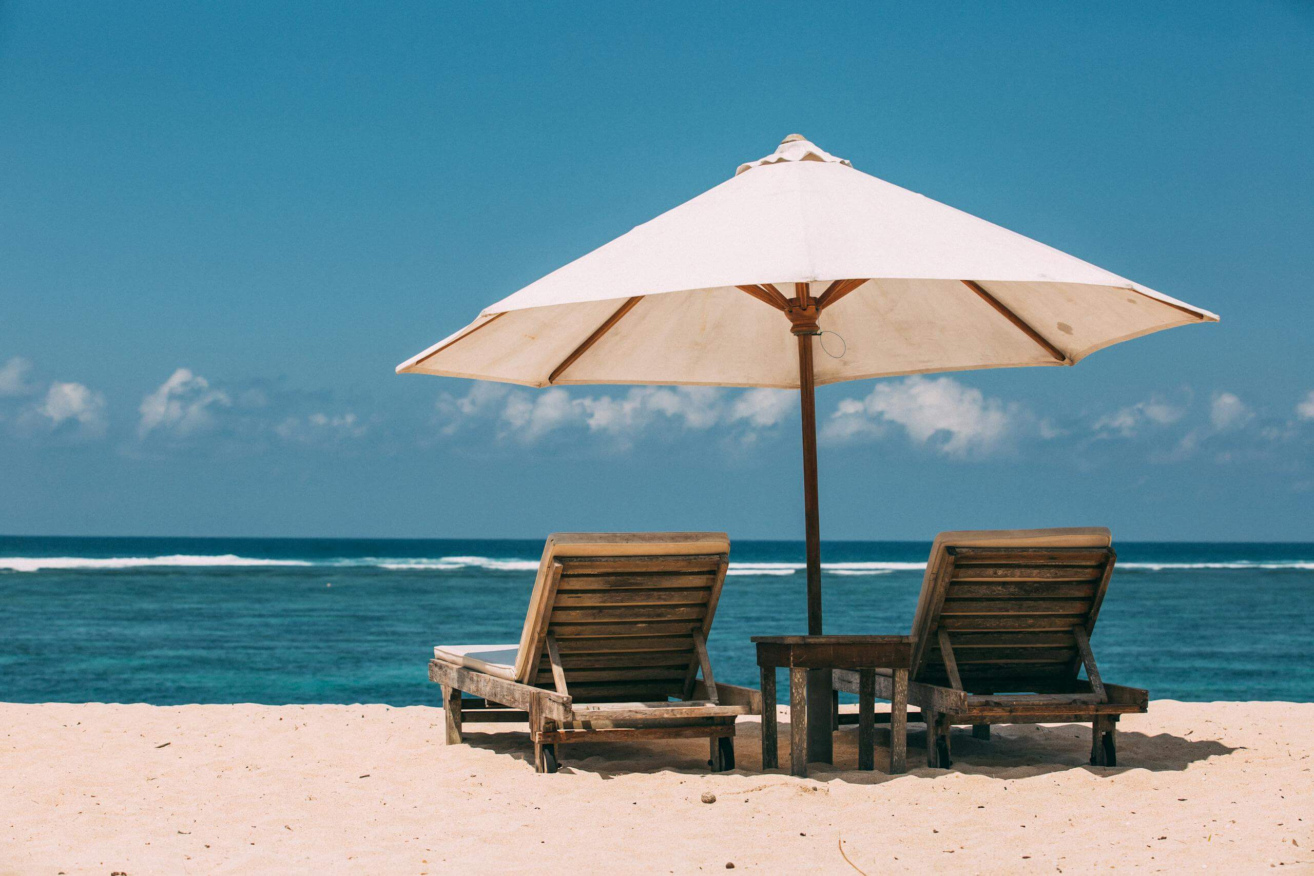 Two sun loungers under an umbrella on a tranquil sandy beach with blue skies.